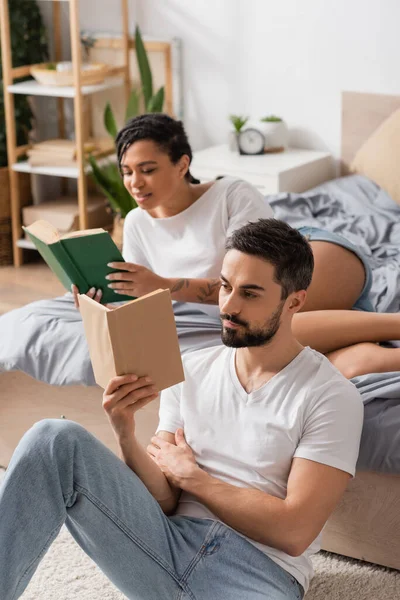 Focalisé homme barbu en jeans lecture livre près flou afro-américaine copine dans la chambre à coucher à la maison — Photo de stock