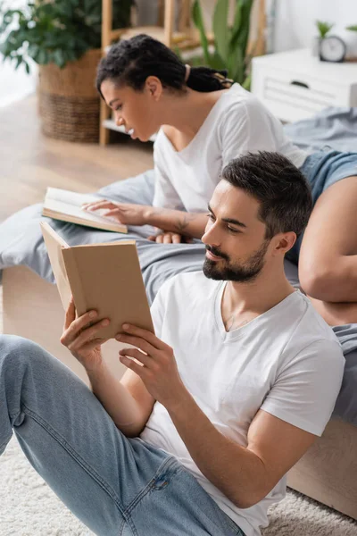 Hombre barbudo positivo y mujer afroamericana joven leyendo libros en el dormitorio en casa - foto de stock