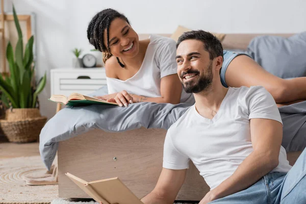 Carefree bearded man sitting with book near young african american girlfriend lying on bed at home — Stock Photo