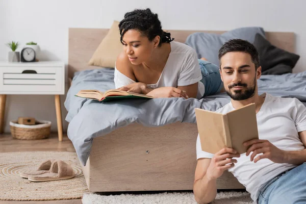 Junge afrikanisch-amerikanische Frau und bärtiger Mann lesen Bücher, während sie sich im Schlafzimmer zu Hause ausruhen — Stockfoto