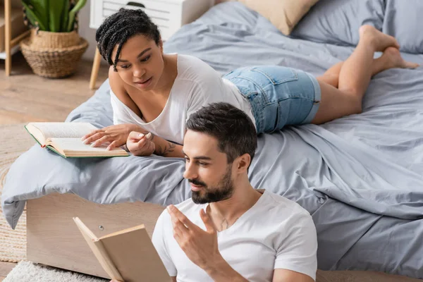 Bearded man gesturing and reading book to african american girlfriend lying on bed at home — Stock Photo