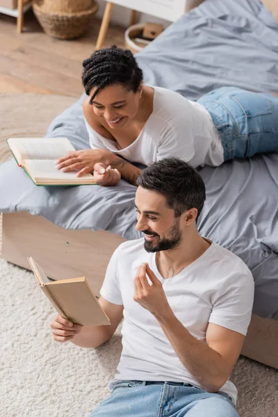 High angle view of cheerful bearded man reading book to smiling african american woman lying on bed at home — Stock Photo