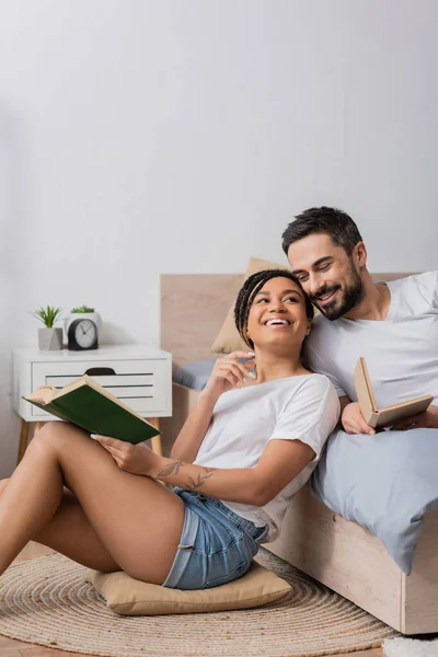 Mulher americana africana alegre com livro sentado no chão e travesseiro perto namorado barbudo deitado na cama em casa — Fotografia de Stock