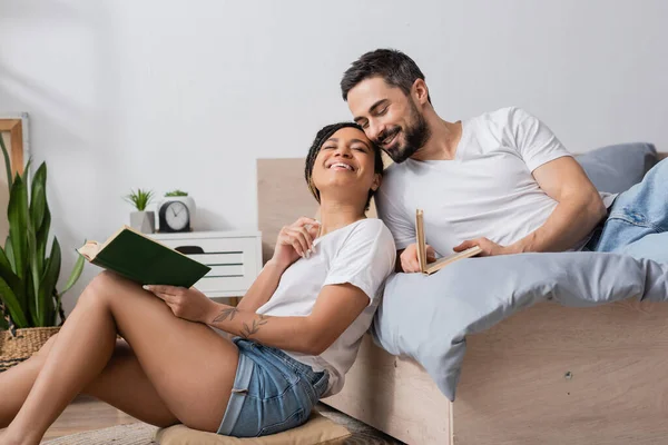 Mulher afro-americana despreocupada com o livro sorrindo com os olhos fechados enquanto sentado perto do namorado sorridente deitado na cama em casa — Fotografia de Stock