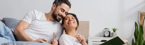 Carefree interracial couple in white t-shirts holding books and smiling with closed eyes in bedroom, banner — Stock Photo