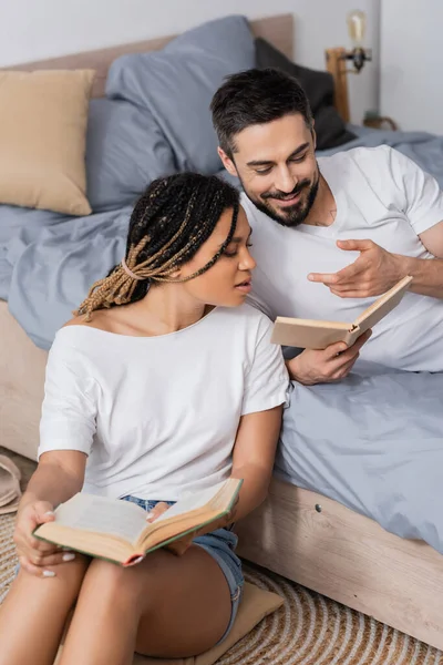 Homme barbu souriant pointant vers livre près de jeune femme afro-américaine dans la chambre à coucher à la maison — Photo de stock