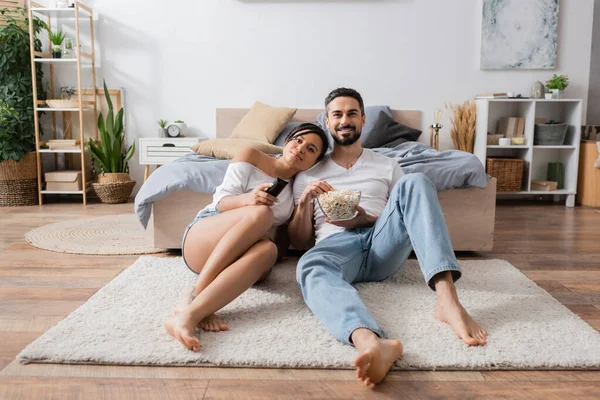 Full length of barefoot and happy interracial couple with bowl of popcorn watching tv on floor in modern bedroom — Stock Photo