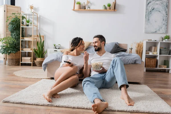 Longitud completa de descalzo y despreocupado pareja interracial sentado con TV mando a distancia y palomitas de maíz en el suelo en el dormitorio moderno - foto de stock