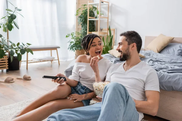 Cheerful bearded man feeding african american woman with popcorn while sitting on floor in modern bedroom — Stock Photo