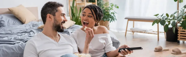 Збуджений бородатий чоловік годує афроамериканку попкорном під час перегляду телевізора в спальні вдома, банер — стокове фото