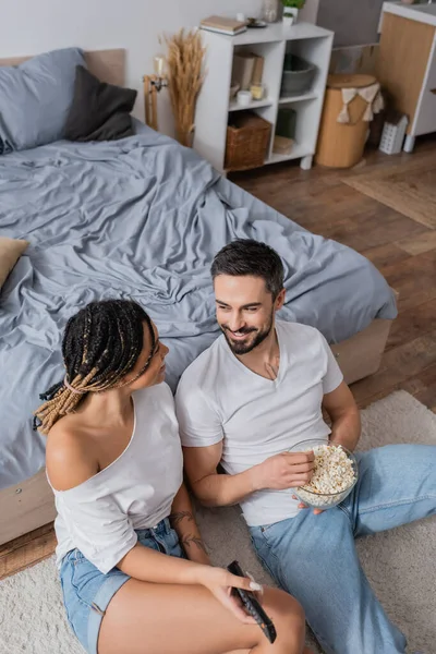 Vue grand angle de couple interracial avec télécommande tv et pop-corn souriant à l'autre sur le sol dans la chambre — Photo de stock