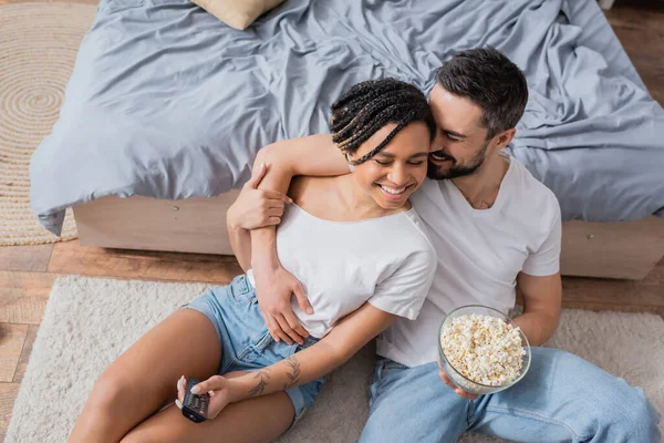 Aus der Vogelperspektive: fröhlicher Mann mit Schüssel Popcorn umarmt afrikanisch-amerikanische Frau mit Fernbedienung auf dem Boden im Schlafzimmer — Stockfoto