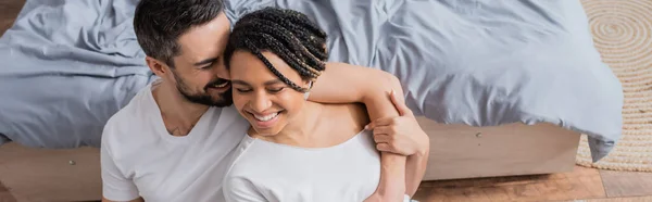 Vista ad alto angolo di uomo barbuto che abbraccia allegra ragazza afroamericana che abbraccia vicino al letto a casa, striscione — Foto stock