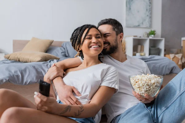 Hombre barbudo con tazón de palomitas de maíz abrazando despreocupado mujer afroamericana con control remoto de televisión en el dormitorio en casa - foto de stock