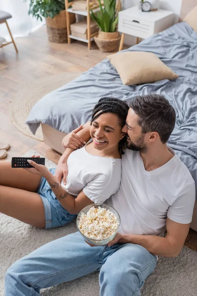 Высокий угол зрения бородатый мужчина с попкорном обнимая радостный африканский американец женщина с телевизором пульт дистанционного управления на полу в спальне — стоковое фото