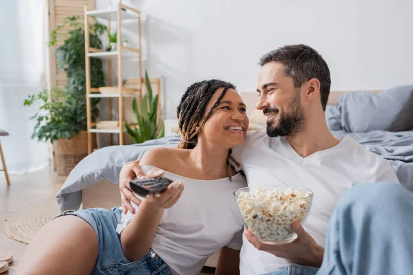 Jeune femme afro-américaine avec télécommande tv et homme barbu avec pop-corn souriant dans la chambre à coucher à la maison — Photo de stock