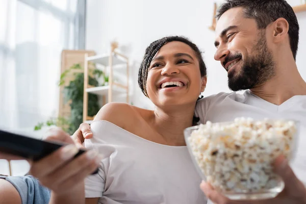 Sonriente hombre barbudo con tazón de palomitas de maíz abrazo despreocupado mujer afroamericana haciendo clic en los canales de televisión en el dormitorio en casa - foto de stock