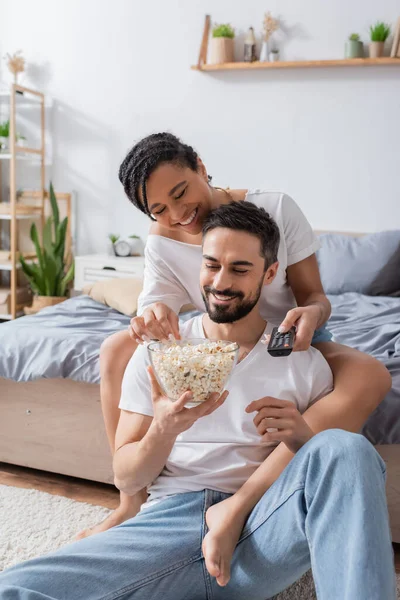 Heureux homme barbu avec bol de maïs soufflé assis sur le sol dans la chambre à coucher près de petite amie afro-américaine avec télécommande tv — Photo de stock