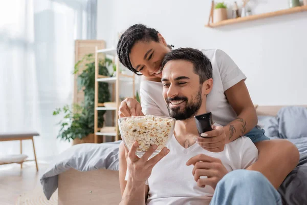 Heureuse femme afro-américaine avec télécommande tv prendre popcorn près de sourire homme barbu dans la chambre à coucher à la maison — Photo de stock