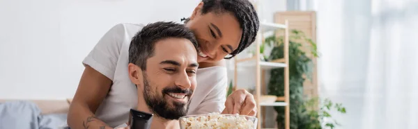 Усміхнений бородатий чоловік тримає попкорн біля безтурботної афроамериканської жінки в спальні вдома, банер — стокове фото