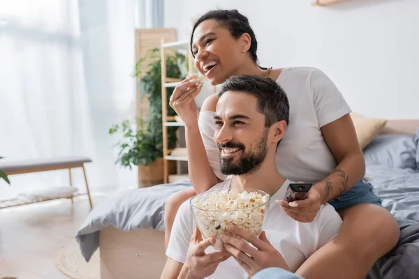 Pleased interracial couple eating popcorn and watching movie in bedroom at home — Stock Photo