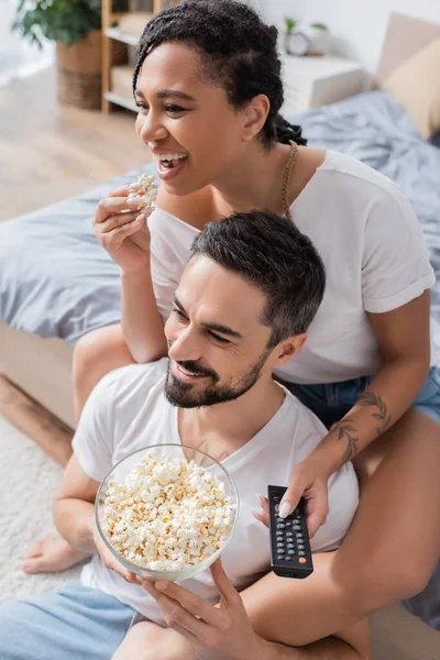 Mujer afroamericana llena de alegría con control remoto de televisión comiendo palomitas de maíz cerca de hombre barbudo sonriente en el dormitorio en casa - foto de stock
