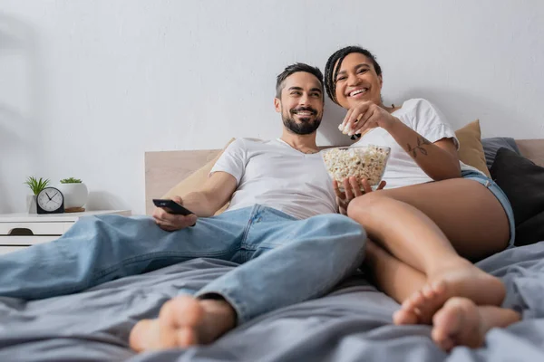 Heureux afro-américaine femme manger pop-corn près sourire homme barbu avec télécommande tv sur le lit à la maison — Photo de stock