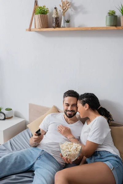 Feliz africano americano mujer abrazando sonriente barbudo hombre con tv control remoto y tazón de palomitas de maíz en la cama en casa - foto de stock
