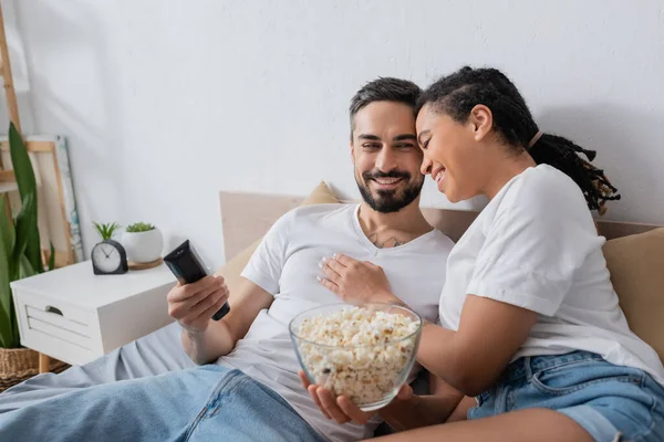 Heureux homme barbu avec bol de maïs soufflé et télécommande tv près de jeune copine afro-américaine sur le lit à la maison — Photo de stock
