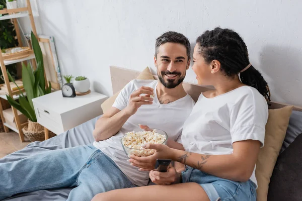 Cheerful interracial couple with tv remote controller and popcorn smiling at each other on bed at home — Stock Photo