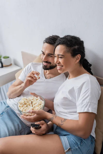 Despreocupados pareja multiétnica en camisetas blancas comiendo palomitas de maíz y viendo la televisión en la cama en casa - foto de stock