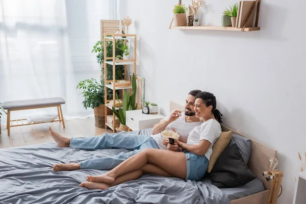 Longitud completa de la pareja interracial descalza comer palomitas de maíz y ver la televisión en el dormitorio espacioso moderno - foto de stock