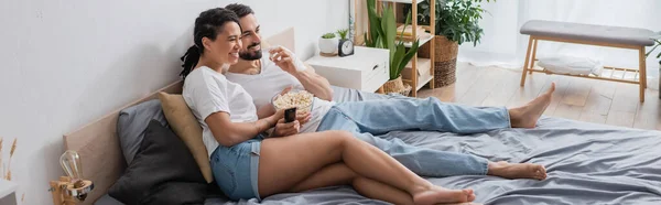 Босоніж і безтурботний міжрасовий пара їсть попкорн і дивиться фільм на ліжку вдома, банер — стокове фото