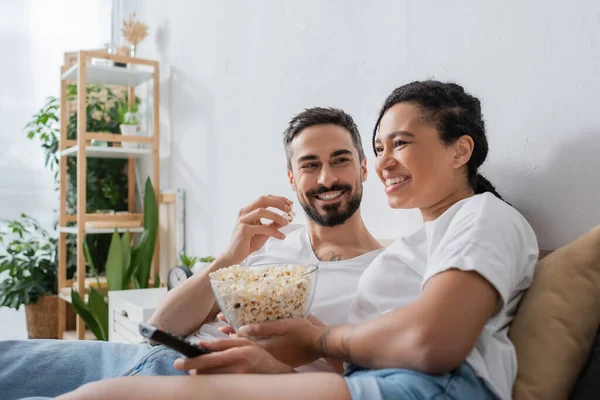 Hombre barbudo complacido comiendo palomitas de maíz y mirando a la sonriente mujer afroamericana con control remoto de televisión en la cama en casa - foto de stock