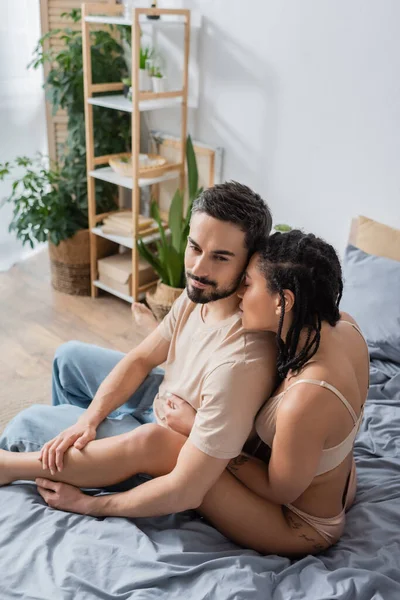 Сексуальная африканская американка в нижнем белье соблазняющая бородатого мужчину сидящего в футболке и джинсах на кровати дома — стоковое фото
