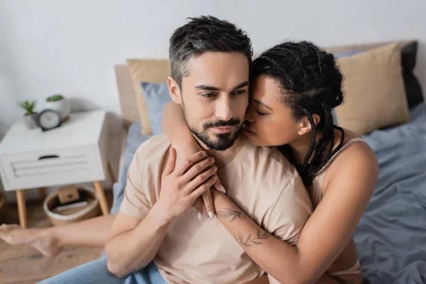 Mujer afroamericana tatuada abrazando a hombre barbudo en camiseta en el dormitorio en casa - foto de stock