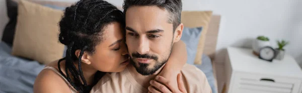 Mujer afroamericana joven con rastas abrazando novio barbudo en el dormitorio en casa, pancarta - foto de stock