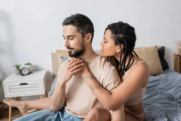Barbudo hombre en camiseta sentado cerca sexy africana americana mujer con rastas en el dormitorio en casa - foto de stock