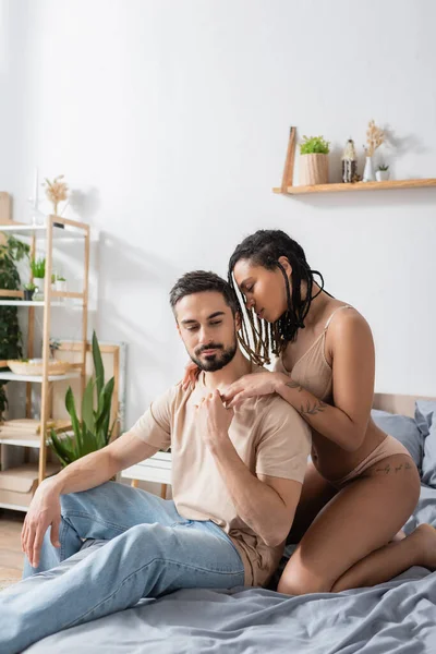 Бородатий чоловік у футболці та джинсах тримає руку татуйованої афроамериканської жінки в нижній білизні в спальні вдома — стокове фото