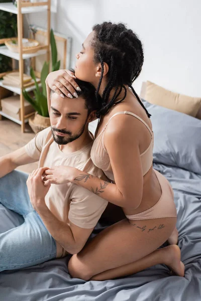 Femme afro-américaine tatouée en lingerie embrassant petit ami barbu assis en t-shirt et jeans sur le lit à la maison — Photo de stock
