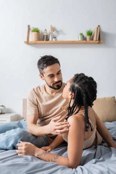 Bärtiger Mann im T-Shirt schaut sexy Afroamerikanerin mit Dreadlocks an, während er zu Hause auf dem Bett sitzt — Stockfoto