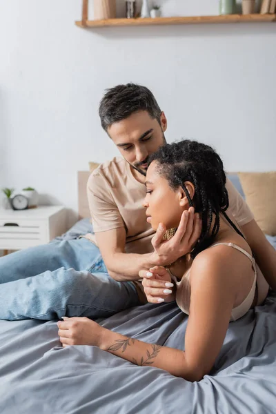 Man in t-shirt and jeans touching face of charming african american woman in lingerie on bed at home — Stock Photo