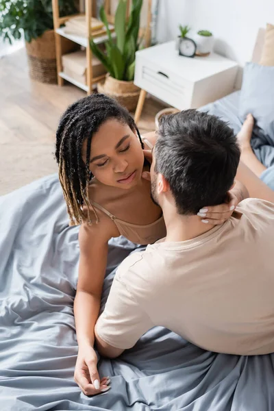 Vista ad alto angolo della donna afroamericana appassionata con gli occhi chiusi vicino all'uomo bruna in t-shirt sul letto a casa — Foto stock