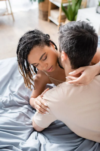 Femme afro-américaine passionnée avec les yeux fermés étreignant homme brune en t-shirt sur le lit à la maison — Photo de stock