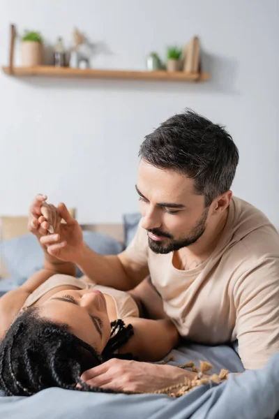 Hombre barbudo en camiseta cogido de la mano de una apasionada mujer afroamericana acostada en la cama en casa - foto de stock