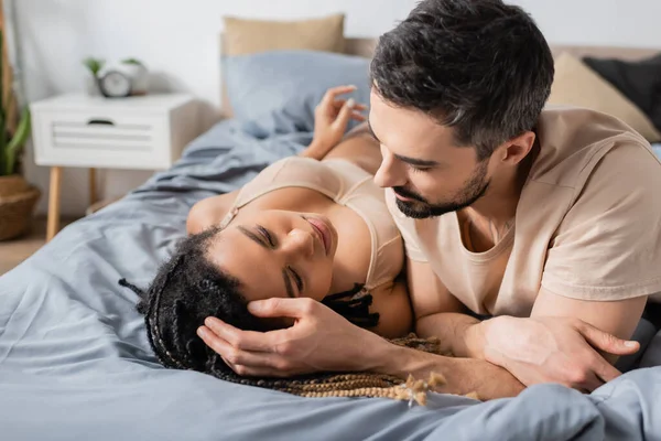 Morena barbudo hombre tocando el pelo de sexy africana americana mujer en lencería acostado en la cama en casa - foto de stock