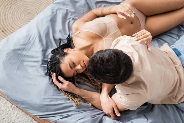 Vista superior de hombre morena tocando el pelo de la mujer afroamericana sexy en ropa interior acostada en la cama con los ojos cerrados - foto de stock