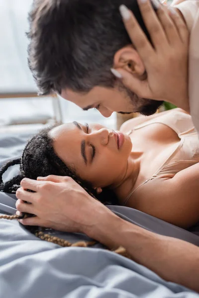 Sexy africana americana mujer en lencería abrazando borrosa hombre en cama en casa - foto de stock