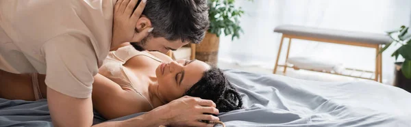 Séduisante femme afro-américaine en soutien-gorge embrassant un homme brune en t-shirt allongé sur le lit à la maison, bannière — Photo de stock
