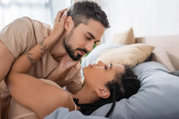 Vista lateral del hombre barbudo en camiseta cerca de la seductora mujer afroamericana acostada en la cama en casa - foto de stock